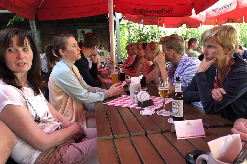 Mitarbeitende sitzen an langen Tischen im Biergarten unter roten Sonnenschirmen und verbringen gemeinsam Zeit bei Getränken und Gesprächen.