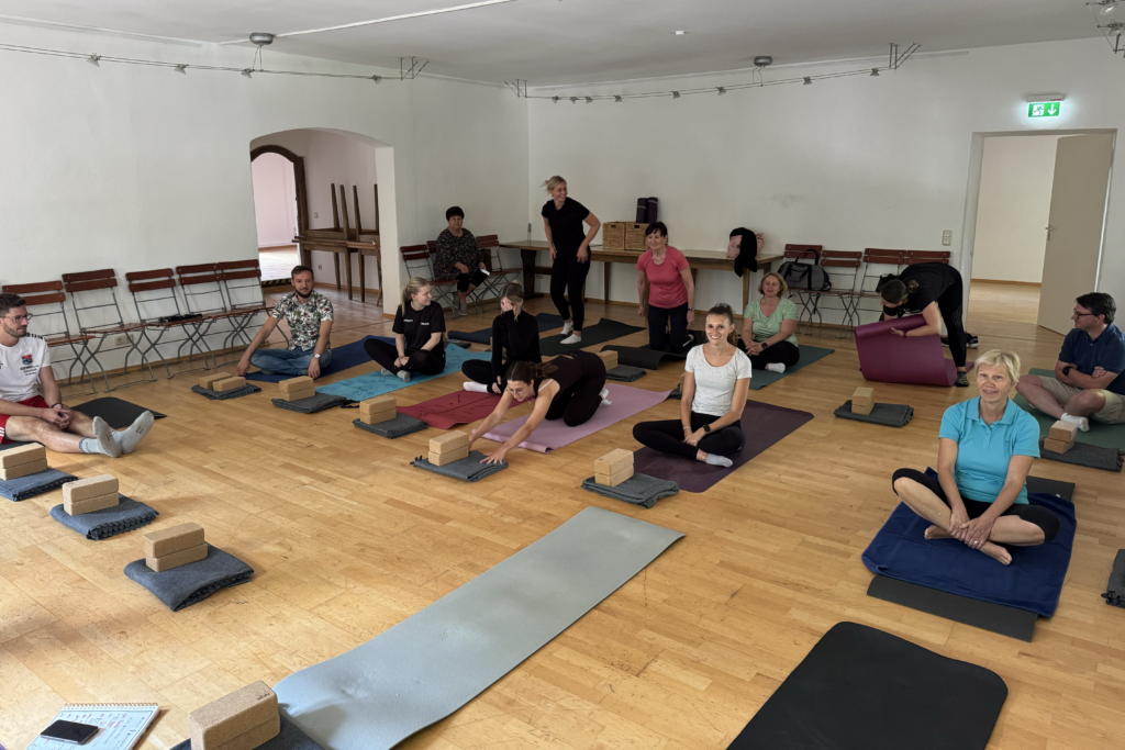 Eine Gruppe von Mitarbeitenden nimmt in einem Kursraum an einer Yoga- bzw. Entspannungseinheit mit Matten und Yoga-Blöcken teil.