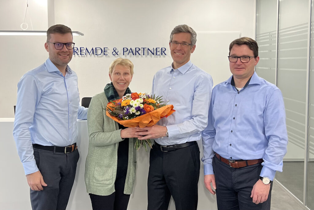 Vier Personen stehen in der Kanzlei vor dem Schriftzug „Remde & Partner“, in der Mitte wird ein bunter Blumenstrauß als Anerkennung überreicht.