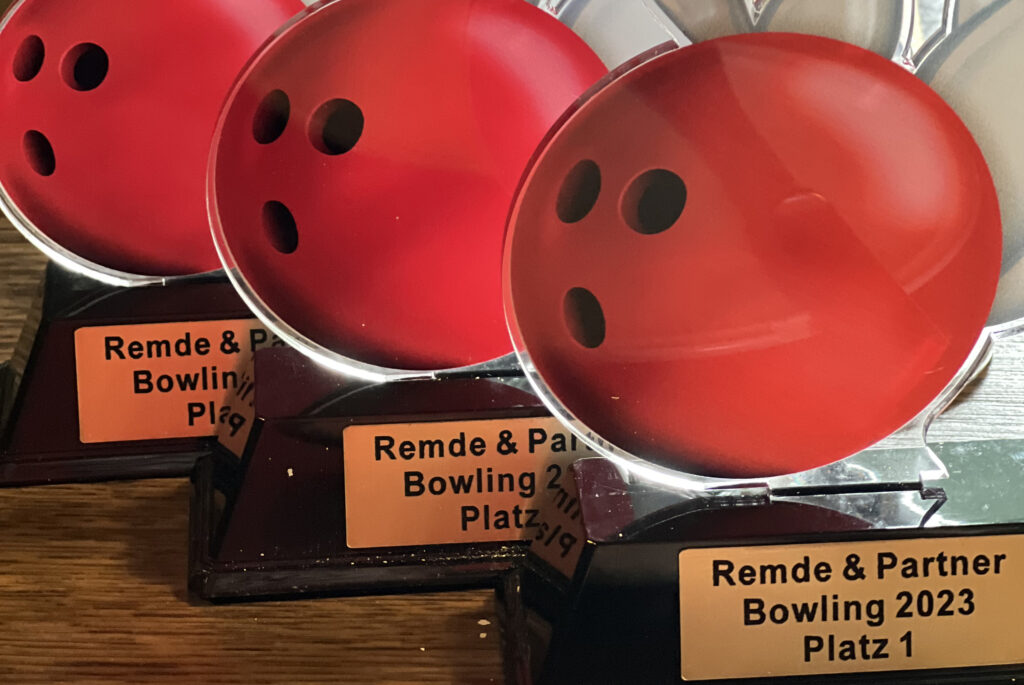 Mehrere Bowling-Pokale mit roten Kugel-Motiven und Gravuren „Remde & Partner Bowling 2023“ für die Plätze 1 bis 3.